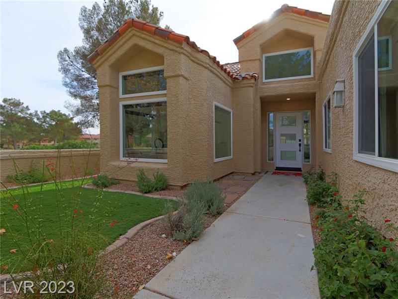 A digital image depicting a modern residential house with a beige stucco finish and terracotta roof tiles. It features windows with white frames, a white front door with a rectangular window, and a concrete path leading to the entrance, bordered by green grass and a landscaped garden with red flowers. The setting has an overcast sky, suggesting a serene suburban environment.