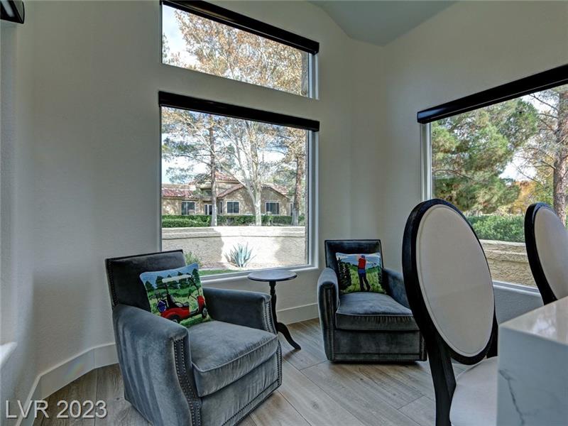 a cozy coffee nook area with two plush armchairs upholstered in a soft gray fabric, each adorned with a colorful cushion featuring a golf motif. Between the chairs is a small, round side table. The area is well-lit by natural light from two large windows with blackout shades, offering a view of a tree-lined golf course and a house across the way. The room has high ceilings and light hardwood floors, creating a bright and inviting space.