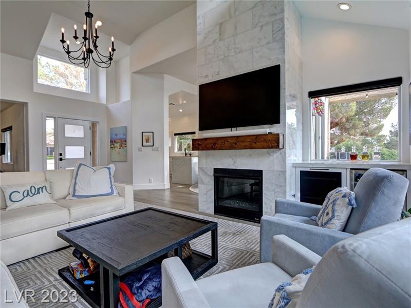 modern living room with a vaulted ceiling and a mix of natural and artificial light. The central feature is a fireplace with a white marble surround and a dark wooden mantelpiece, above which hangs a flat-screen TV. A large, comfortable white sofa with 'calm' embroidered on a cushion, and a gray armchair with a blue throw blanket, provide seating around a dark wooden coffee table with storage underneath. On the right, a small wine fridge is built into the white cabinetry. The room's decor is completed with geometric-patterned gray area rugs on light hardwood floors, and a simple black chandelier adds a touch of elegance