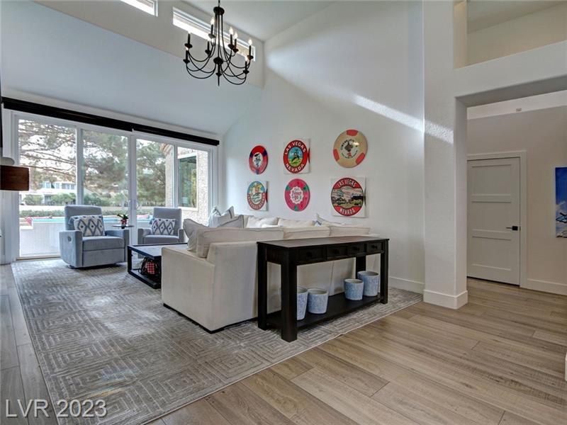 n interior photograph of a spacious living room featuring high ceilings and large windows allowing for ample natural light. The room is furnished with a white sofa facing two patterned armchairs, a dark wooden dining table with white tablecloth, and a sideboard against the wall. The floor is covered with a gray patterned area rug over light wooden flooring. On the wall, decorative plates with vibrant designs add a pop of color. The room's color palette is a mix of neutrals with accent colors from the wall art and blue patterned ceramics.