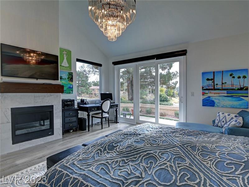 spacious primary bedroom featuring a large bed with an intricate blue and white patterned comforter. Above the bed hangs an elegant crystal chandelier. To the left is a cozy workspace with a black desk and chair, a green hourglass artwork above the desk, and a fireplace with a marble surround similar to the one in the living room. The room has large sliding glass doors leading to a balcony with a view of pine trees and a clear sky. On the opposite wall is a colorful painting depicting a beach scene. The room combines a serene color palette with vibrant art accents and ample natural light, creating a tranquil yet lively atmosphere.