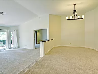 The image shows a spacious, light-filled interior with an open layout. The room features soft beige carpeting and neutral-colored walls, creating a warm and inviting atmosphere. A stylish chandelier hangs from the ceiling, providing elegant lighting to the space. The area is divided into a dining section and a living area, with a partial wall separating the two spaces. In the background, large windows and glass doors lead to the outdoor patio, allowing plenty of natural light to enter and offering views of the greenery outside.