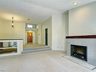The image showcases a cozy living area with a modern fireplace as a focal point. The fireplace features a sleek design with a tile surround and a wooden mantle. The room is carpeted and painted in soft, neutral colors, enhancing the bright and airy feel of the space. An elegant light fixture is visible in the background, illuminating the dining area. The room has a split-level design, with a few steps leading up to a hallway and additional living spaces. A large mirror on the half-wall adds depth and reflects light, contributing to the open and inviting ambiance.