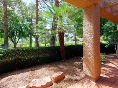 The image shows a peaceful backyard space featuring a small garden area with a palm tree and other greenery. The garden is enclosed by a low, green hedge, providing a sense of privacy. A tiled patio area extends from the home, shaded by an overhanging pergola. The view beyond the garden reveals tall trees and a well-maintained lawn, creating a tranquil and inviting outdoor setting.