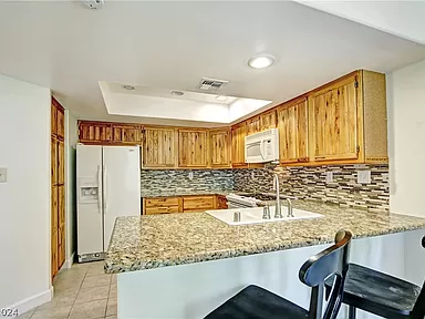 The image depicts a modern kitchen with a warm, inviting design. The kitchen features granite countertops and a backsplash with a mix of neutral-toned tiles. The wooden cabinets provide ample storage and add a rustic charm to the space. A white refrigerator and microwave complement the clean, bright look of the kitchen. A dual-basin sink is situated in the counter, which also serves as a breakfast bar with two black chairs for seating. The room is well-lit with recessed lighting and natural light from the surrounding area.