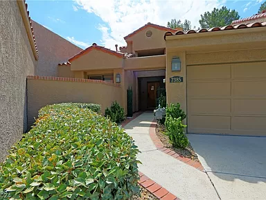 The image shows the front exterior of a single-story townhome located at 7385 Mission Hills Drive. The house features a beige stucco finish with red-tiled roof accents. The entrance is framed by a small, well-maintained garden with green shrubs. A paved pathway leads to the front door, flanked by two potted plants, and a single-car garage is visible on the right. The overall appearance is welcoming and well-kept, set against a backdrop of a partly cloudy sky and mature trees.