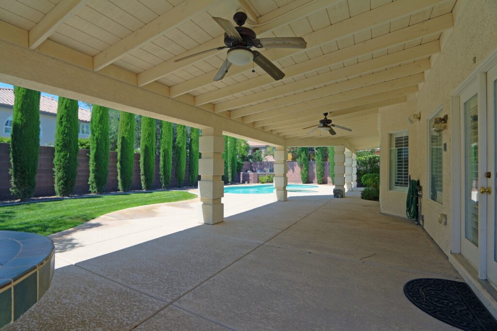 A spacious covered patio with ceiling fans, overlooking a backyard with a swimming pool, lush green lawn, and tall cypress trees providing privacy.