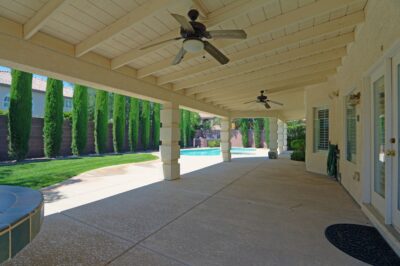A spacious covered patio with ceiling fans, overlooking a backyard with a swimming pool, lush green lawn, and tall cypress trees providing privacy.