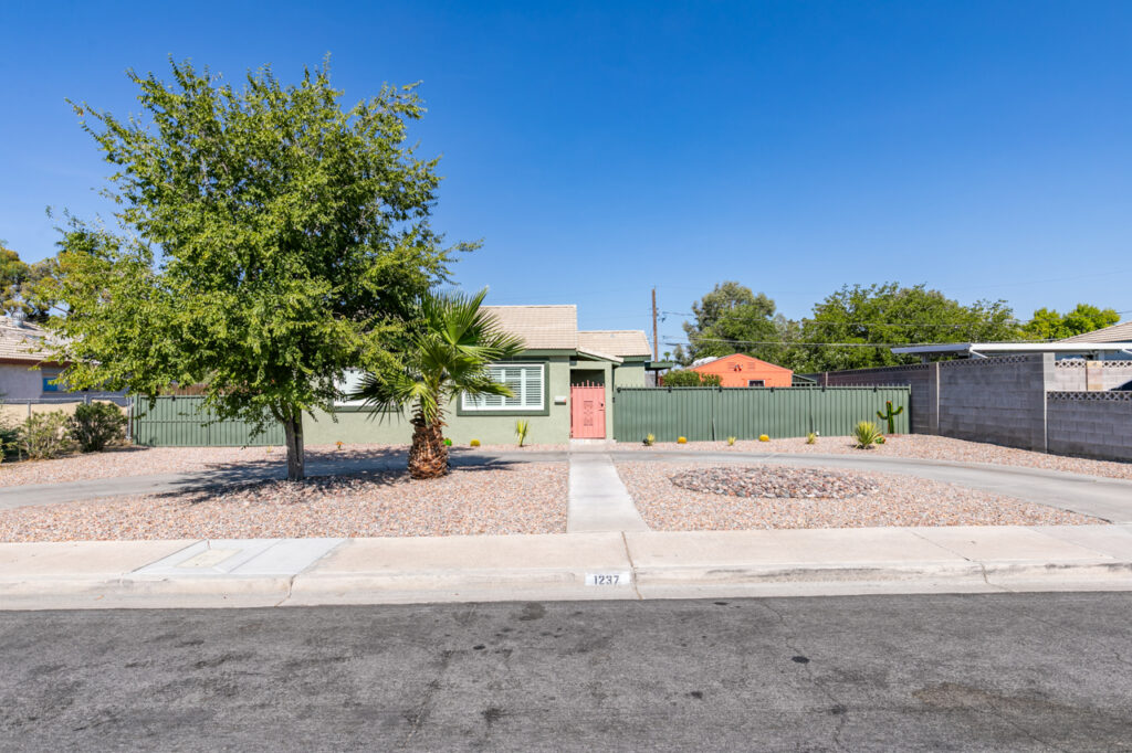 A single-story house with a light green exterior and a red front door, surrounded by a low green fence. The front yard features a large tree, a small palm tree, and a minimalist desert landscape with gravel and a circular rock arrangement. The house is set against a bright blue sky.