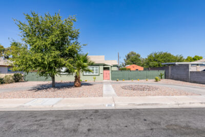 A single-story house with a light green exterior and a red front door, surrounded by a low green fence. The front yard features a large tree, a small palm tree, and a minimalist desert landscape with gravel and a circular rock arrangement. The house is set against a bright blue sky.