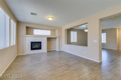 A spacious living room with beige walls, wood-look tile flooring, a built-in fireplace with a white tile surround, and an open layout with cut-out wall openings leading to adjacent spaces.