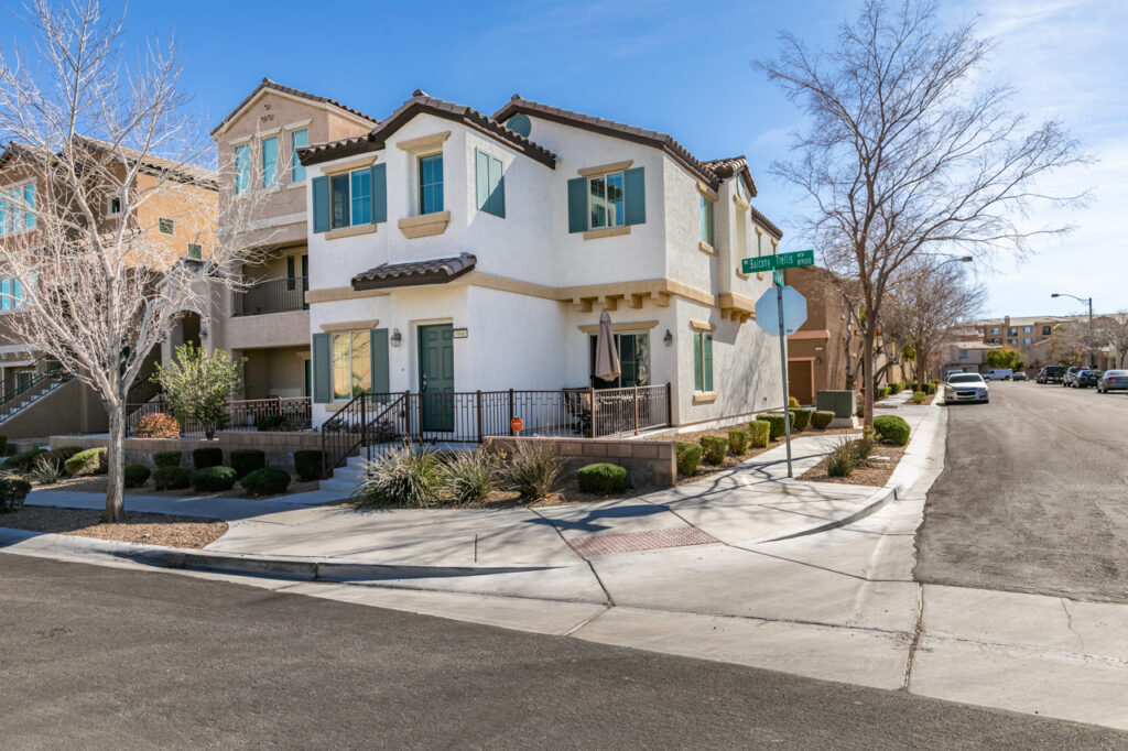 A two-story beige and white house with brown shutters and a brown garage door sits on a corner lot. A stop sign and street signs are visible at the intersection, with a clear blue sky and trees in the background. The driveway is spacious, and neighboring homes can be seen nearby.