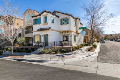 A two-story beige and white house with brown shutters and a brown garage door sits on a corner lot. A stop sign and street signs are visible at the intersection, with a clear blue sky and trees in the background. The driveway is spacious, and neighboring homes can be seen nearby.