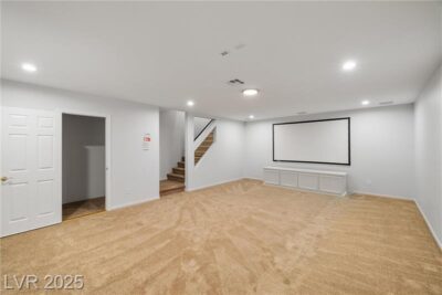 A spacious basement with beige carpeting, white walls, recessed lighting, and a built-in projector screen with a media cabinet below. A staircase leads to the upper level, and there is an open storage area to the left.