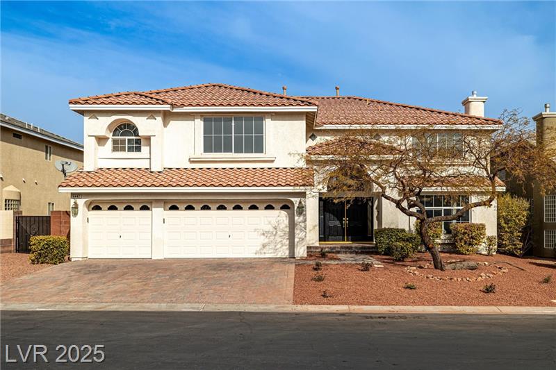 A two-story Mediterranean-style home with a three-car garage, a tile roof, and a desert-landscaped front yard. The house features large windows and an inviting entryway with a glass front door. The sky is clear and blue, highlighting the home's bright exterior.