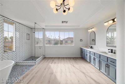 A spacious primary bathroom with a large walk-in shower featuring herringbone tile, a freestanding soaking tub, a double vanity with blue cabinetry, and modern lighting fixtures. Large windows provide natural light and a view of neighboring homes.