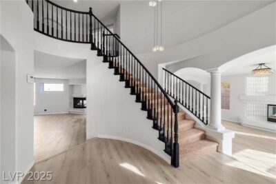 A bright and spacious living room with light wood flooring, white walls, and elegant arched columns. The room features a modern fireplace with a marble surround, large windows allowing natural light to flood in, and a stylish chandelier hanging from the ceiling. A grand staircase with wrought iron railings leads to the upper level.