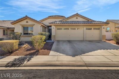 A single-story suburban home with a three-car garage, a beige stucco exterior, and a desert-landscaped front yard with trimmed bushes and rocks. The house has a tiled roof and a concrete driveway leading to the garage.