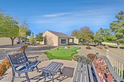 A beautifully landscaped backyard featuring a private putting green, desert-friendly landscaping, and a cozy seating area with decorative metal benches. The home has a covered patio and is set against a scenic backdrop with mature trees and a golf course view.