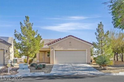 A single-story, beige stucco home with a two-car garage and desert landscaping in a suburban neighborhood. The front yard features drought-tolerant plants, trees, and a paved driveway leading to the garage. A clear blue sky is visible above.