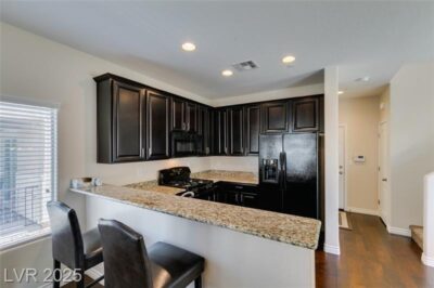 A modern kitchen with dark cabinetry, granite countertops, and stainless steel appliances, including a black refrigerator and gas stove. The kitchen features a breakfast bar with two black barstools, recessed lighting, and an open layout connecting to the living space.