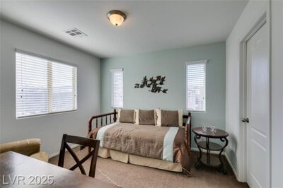 A well-lit bedroom featuring a wooden daybed with brown and light blue bedding, three decorative pillows, and a metal bird wall decoration above the bed. The walls are painted light blue and white, with two small windows flanking the bed. A round wooden side table with a glass top and two small glasses is positioned next to the bed. A wooden desk and chair are in the foreground, with a beige upholstered chair partially visible. The ceiling has a simple light fixture, and natural light streams through the windows.