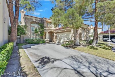 A stunning two-story home with a Mediterranean-style design, featuring a beige stucco exterior, red tile roof, and large arched windows. The home is surrounded by lush greenery and mature trees, with a well-maintained front yard and a spacious driveway leading to a two-car garage.