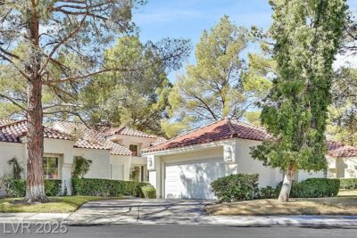 A beautiful single-story home with a Mediterranean-style design, featuring a white stucco exterior, red tile roof, and lush green landscaping. A two-car garage with a white door is visible, along with tall trees and well-manicured bushes lining the front yard. The home is situated in a peaceful neighborhood with bright blue skies overhead.