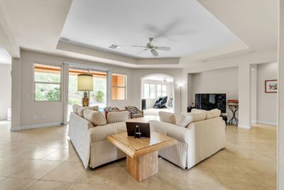 Bright and spacious living room with tile flooring, neutral-toned furniture, tray ceiling, and large windows offering backyard views.