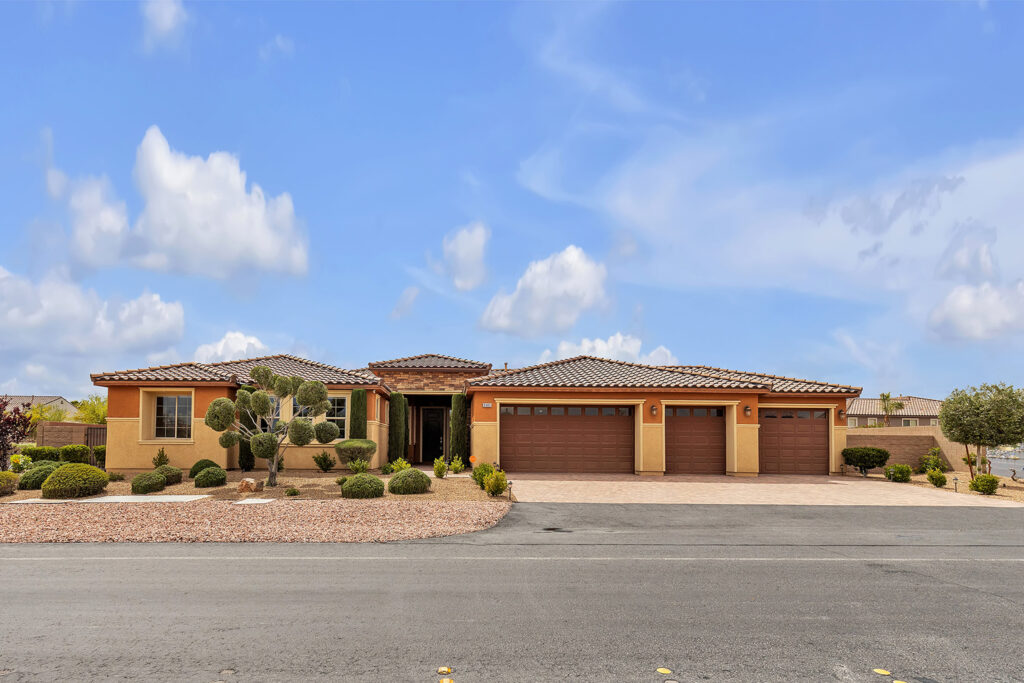 Front exterior of a single-story home with a tile roof, four-car garage, desert landscaping, and a clean, modern stucco finish under a bright blue sky.