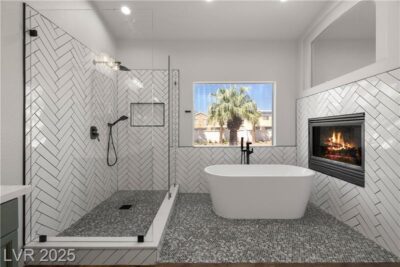 Modern bathroom with glass walk-in shower, freestanding tub, herringbone tile walls, and built-in fireplace beneath a large window.