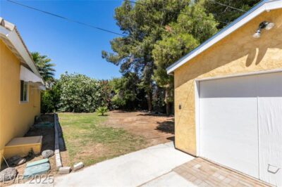 Backyard with mature trees, partial lawn, and a detached yellow garage under a clear blue sky.