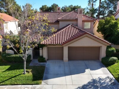 Front exterior view of a two-story home for sale in Spanish Trail, Las Vegas featuring a red tile roof, three-car garage, well-manicured lawn, mature shade trees, and lush landscaping. Located in a guard-gated golf community and includes access to a private backyard pool